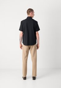 Cămașă neagră cu mâneci scurte și guler mandarin, asortată cu pantaloni bej deschis. Cămașa are o textură netedă și un tiv curbat.