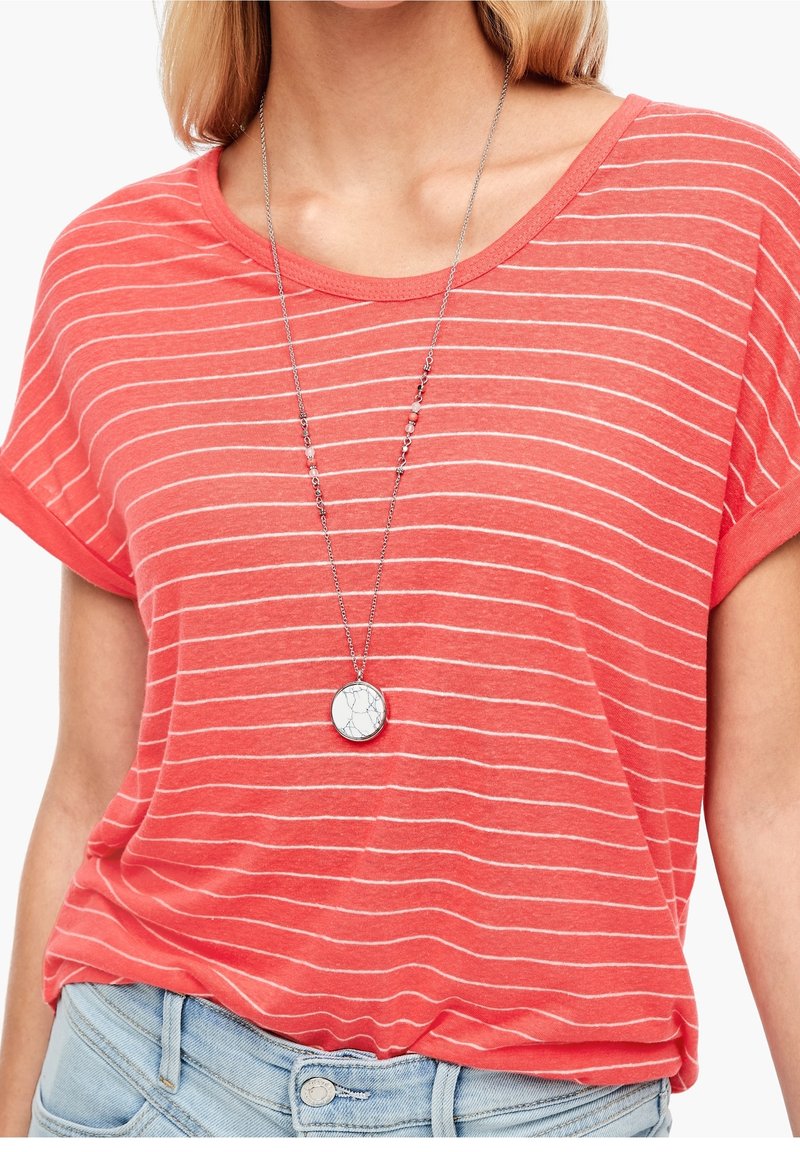 Femme portant un t-shirt rayé rouge et blanc à manches courtes, un jean bleu clair et un long collier argenté avec un pendentif rond blanc.