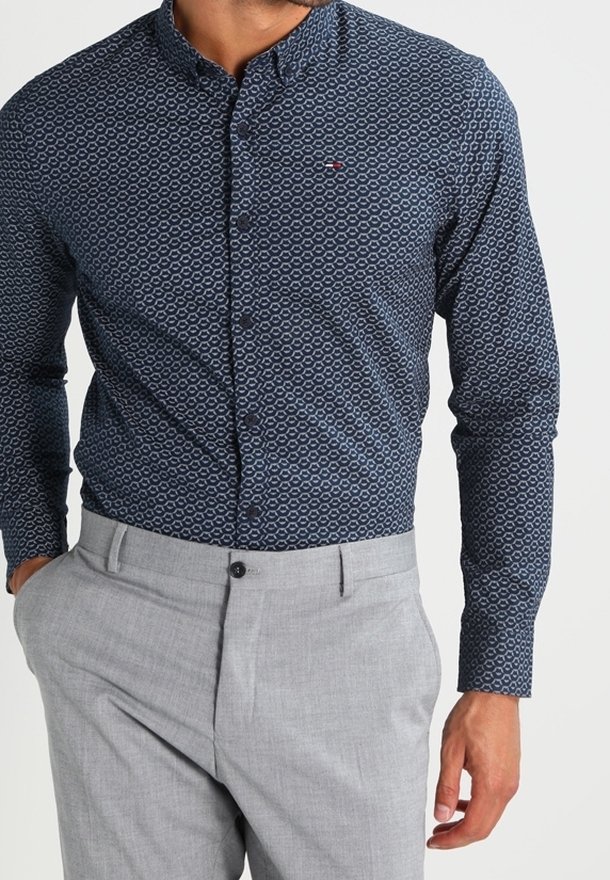 Homme portant une chemise bleu foncé à motifs avec manches longues et un pantalon habillé gris clair, main gauche dans la poche, face à l’avant.
