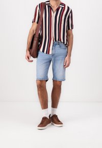 Man wearing multi-color vertical striped short-sleeve shirt, light blue denim shorts, white socks, brown sneakers, and carrying a brown shoulder bag.