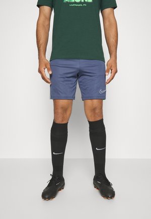 Athlet trägt grünes Liverpool FC Trikot, blaue Nike-Shorts, schwarze Nike-Socken und schwarze Fußballschuhe und steht auf grauem Boden.