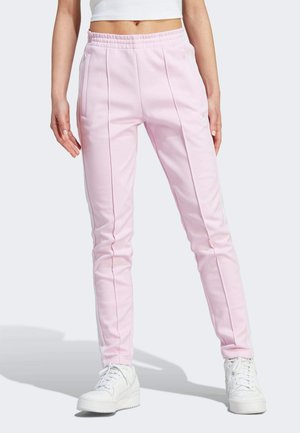 Kvinde iført lyserøde, smalle træningsbukser med hvide sidelommer, hvide sneakers og en hvid crop top, stående med afslappede arme.