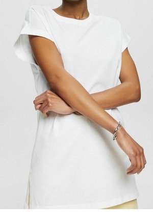 Persona con camiseta blanca de manga corta con puños remangados, brazos cruzados y una pulsera plateada en la muñeca derecha sobre un fondo liso.