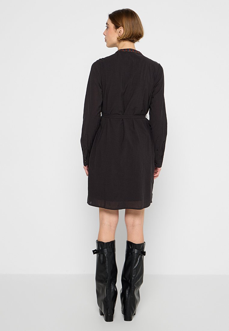 Schwarzes Kleid mit langen Ärmeln und einem Bindegürtel, mit einer Knopfverschluss an den Bündchen und einem knielangen Saum. Getragen mit schwarzen Stiefeln bis zum Knie.