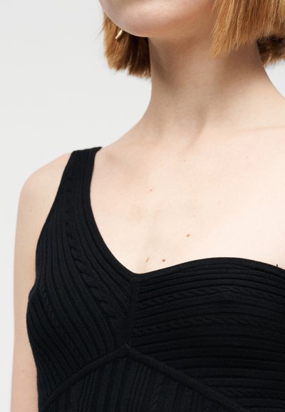 Débardeur en maille côtelée noire avec un col en V, présentant des motifs torsadés texturés et des bords lisses. Design simple, tissu léger.