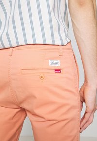 Homme portant un pantalon Levi's couleur pêche avec une poche arrière boutonnée, une étiquette Levi's blanche et une chemise rayée rentrée.