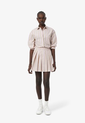 Frau steht und trägt ein beige-weiß gestreiftes Hemd mit senkrechten Streifen, mit puffigen Ärmeln, einen passenden plissierten Rock, weiße Socken und weiße Turnschuhe.