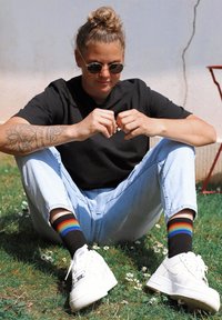 Zapatillas blancas, pantalones vaqueros de un azul claro, camiseta negra y calcetines a rayas de colores. Modelo sentado en la hierba con pequeñas flores, tatuajes visibles en el brazo.
