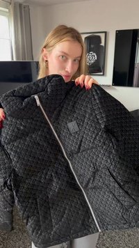 Mladá žena s blond vlasmi drží čiernu prešívanú bundu so strieborným zipsom a jemným opakujúcim sa vzorom v interiéri.