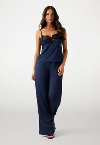 Femme portant un débardeur en satin bleu marine avec une dentelle noire et un pantalon large en satin assorti, marchant vers l'avant sur un fond clair.