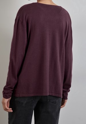 Burgundroter Oversized-Pullover mit langen Ärmeln und einem weiten Rundhalsausschnitt. Weiche Textur, leicht verlängert hinten, kombiniert mit dunklen Jeans.