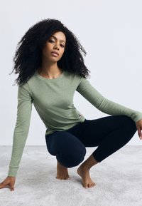 Langarmige groene ribgebreide top met ronde hals, gecombineerd met donkere leggings. Opvallend logo-accent aan de zijkant en krullend haar in het zicht.