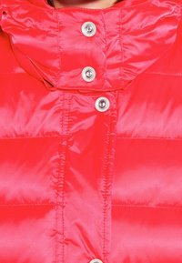 Veste matelassée rouge avec un col haut, dotée de boutons-pression et d'une texture brillante et lisse. Design matelassé avec des coutures visibles.