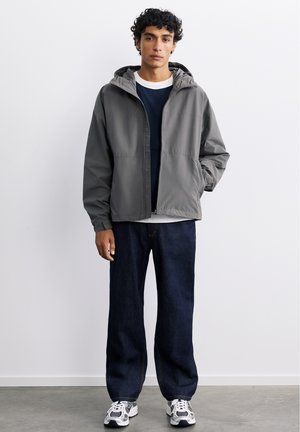 Ung mand med krøllet hår iført en grå hættetrøje, mørke jeans, hvide og sorte sneakers, stående med den ene hånd i lommen.
