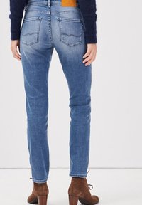 Jeans de mezclilla azul con un diseño ajustado, bolsillos traseros con una costura curva y una etiqueta de cuero marrón en la cintura. Botines marrones visibles.