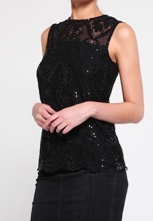 Musta hihaton paita, jossa on pitsikerros ja paljettikoristelu, aaltoileva helma ja korkea kaulus, yhdistetty mustaan farkkuhameeseen.