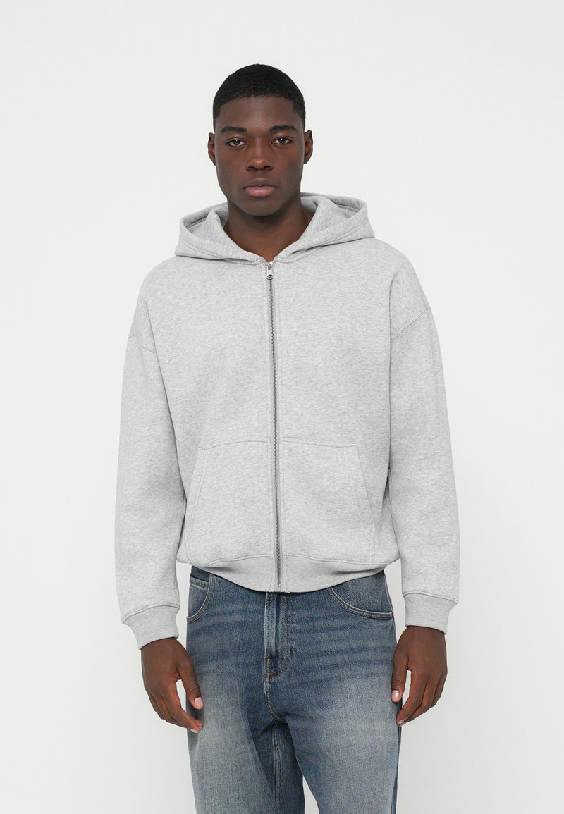 Jauns vīrietis, kurš ģērbts gaiši pelēkā kapucē ar rāvējslēdzēju un zilos džinsos, stāv pretī vienkāršam baltam fonam.