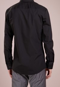 Mann in schwarzem Langarmshirt und grauer Hose, von hinten vor neutralem Hintergrund zu sehen.