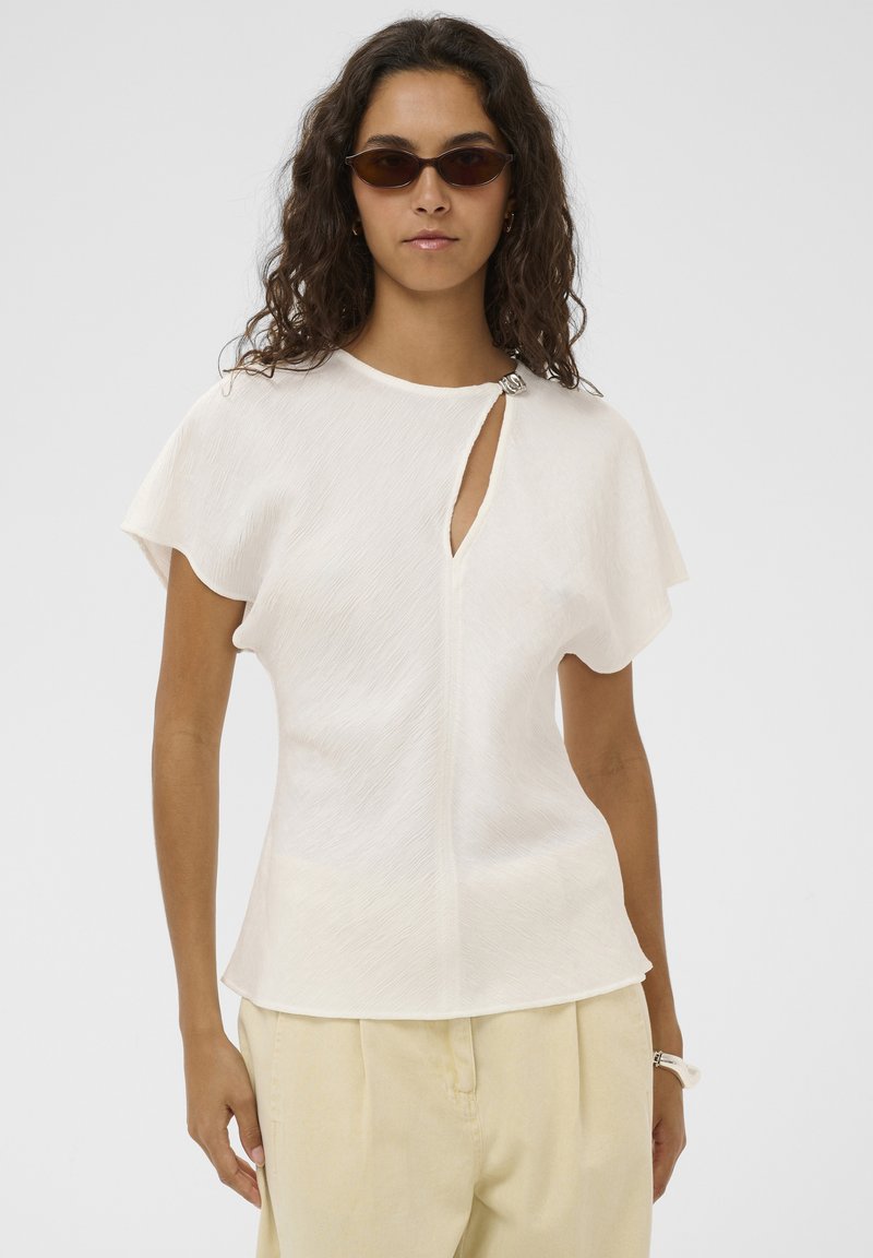 Femme portant un haut blanc à manches courtes avec fermoir à l'avant et détail en forme de trou de serrure, pantalon beige, lunettes de soleil foncées et cheveux bouclés détachés.