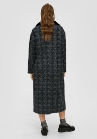 Langer, dunkel gemusterter Mantel mit Karodesign, schwarzem Kragen und Seitentaschen. Getragen über schwarzen Plateauschuhen und gemusterten Socken.