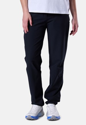 Personne portant un pantalon noir à jambes droites, des chaussures de sport blanches avec des accents bleus, et une chemise blanche, debout les pieds à plat sur le sol.