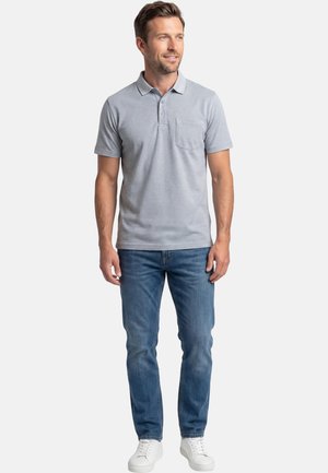 Mies vaaleanharmaassa poolopaidassa, jossa on tasku, sinisissä farkuissa ja valkoisissa lenkkareissa, seisoo ja katsoo hieman oikealle.