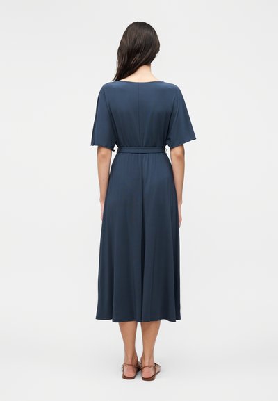 Femme aux longs cheveux foncés, debout, vue de dos, portant une robe midi bleu foncé à manches courtes et des sandales plates marron.