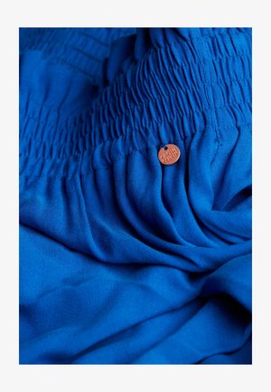 Tissu bleu avec une texture douce, comportant des sections froncées et une petite étiquette ronde avec le nom de la marque.