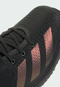 Zbliżenie czarnego buta sportowego z trzema odblaskowymi miedzianymi paskami i czarnymi sznurowadłami na teksturowanej górze z tkaniny.