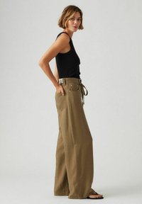Bredde ben olivenbukser laget av teksturert stoff, med høy midje og knyttet belte. Kombinert med en tettsittende svart tanktop og sandaler.