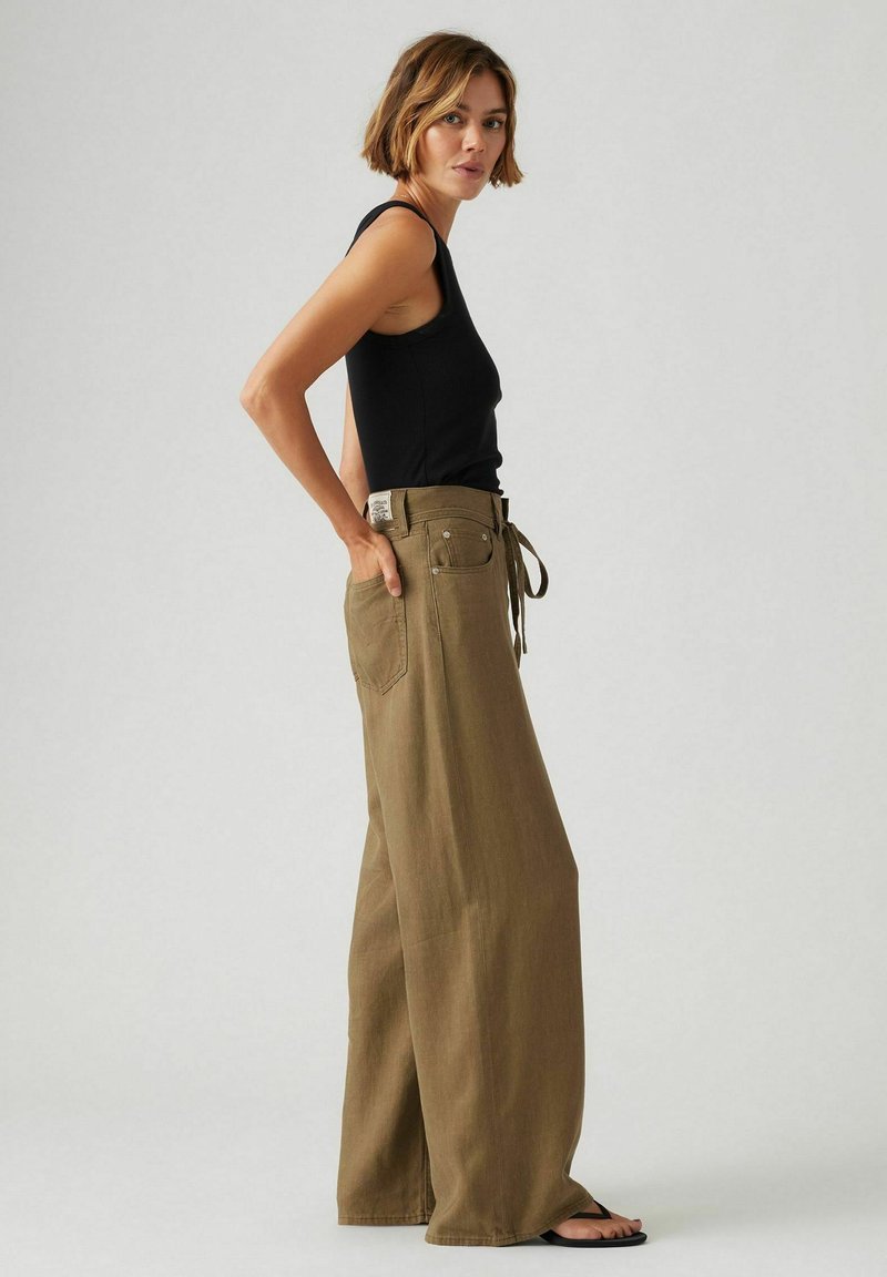 Pantalons olive à jambe large en tissu texturé, avec une taille haute et une ceinture nouée. Associés à un débardeur noir ajusté et des sandales.