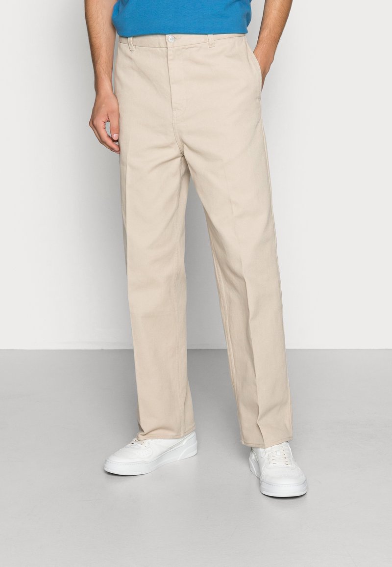 Pantaloni beige con vestibilità dritta, realizzati in tessuto di cotone. Presentano una chiusura con bottone e tasche laterali. Abbinati a sneakers bianche.