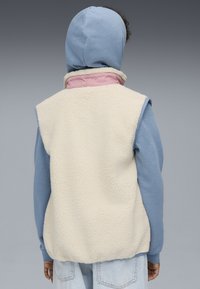 Fleece-vestti off-white värissä, jossa on korkea kaulus ja vaaleanpunaiset yksityiskohdat, päällä sininen pitkähihainen huppari. Rakenne on teksturoitu ja ilman hihoja.