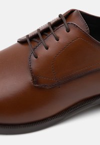 Zapato de vestir de cuero marrón con textura suave, puntera redonda, cierre de cordones, costuras en tono y suela de goma negra.