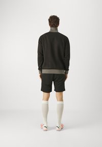 Giacca atletica nera con colletto alto, abbinata a pantaloni corti neri e calze bianche fino al ginocchio. Scarpe da ginnastica con accenti rosa brillante visibili.