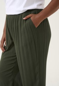 Vihreät laskostetut housut, jotka on valmistettu kevyestä kankaasta, ja joissa on joustava vyötärö ja sivutaskut toiminnallisuuden vuoksi.