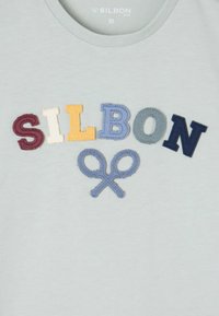 Camiseta de algodón azul claro con letras bordadas de varios colores que dicen "SILBON" y un diseño de tijeras azules debajo.