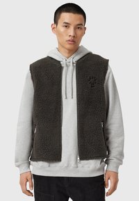 Gilet en polaire noir avec poches zippées, porté sur un sweat à capuche gris. Présente une surface texturée et un design à ouverture frontale. Détail de logo minimaliste visible.