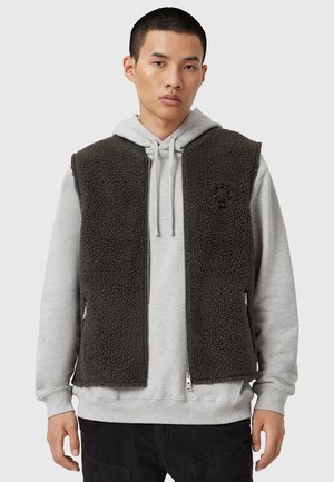Gilet en polaire noir avec poches zippées, porté sur un sweat à capuche gris. Présente une surface texturée et un design à ouverture frontale. Détail de logo minimaliste visible.