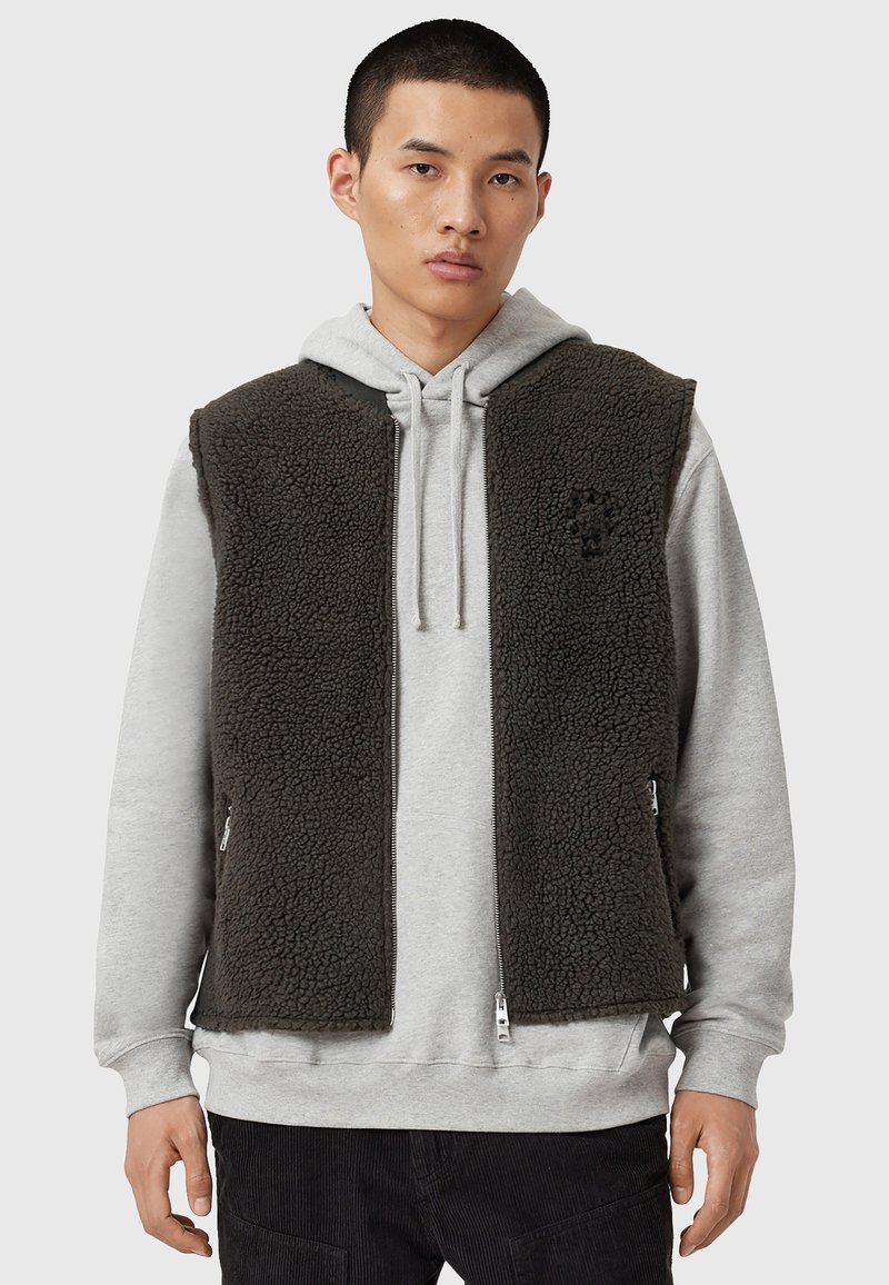 Gilet en polaire noir avec poches zippées, porté sur un sweat à capuche gris. Présente une surface texturée et un design à ouverture frontale. Détail de logo minimaliste visible.