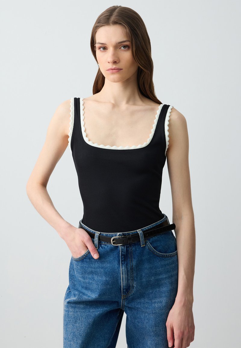 Body nero a costine con scollatura quadrata, dotato di bordo scallop in crema. Abbinato a jeans in denim blu a vita alta e una cintura nera.