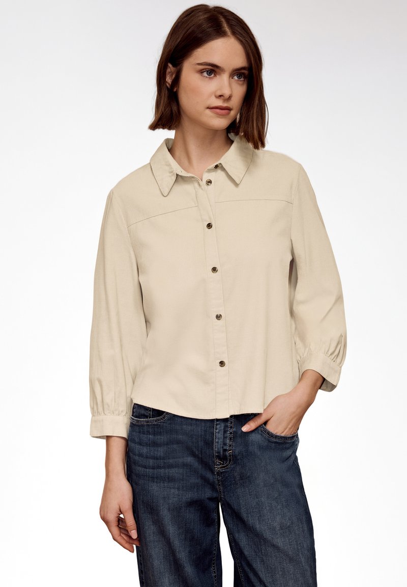 Jeune femme portant une chemise beige à boutons avec des manches trois-quarts et un jean bleu foncé, debout avec une main dans la poche.