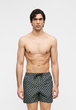 Homme torse nu aux cheveux bouclés foncés, portant un short de bain noir avec un motif de vagues blanches répétées, debout devant un fond clair uni.