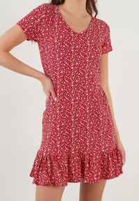 Rotes Blumenkleid mit V-Ausschnitt, kurzen Ärmeln und einem gerafften Saum. Der Stoff ist mit einem kleinen weißen Blumenmuster versehen.