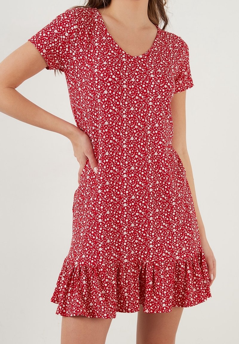 Rotes Blumenkleid mit V-Ausschnitt, kurzen Ärmeln und einem gerafften Saum. Der Stoff ist mit einem kleinen weißen Blumenmuster versehen.