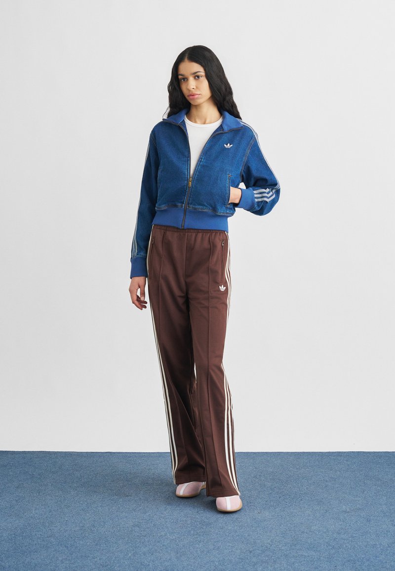 Chaqueta azul de denim con cremallera recortada y rayas blancas, combinada con pantalones deportivos marrones con rayas blancas en los laterales. Calzado rosa completa el conjunto.