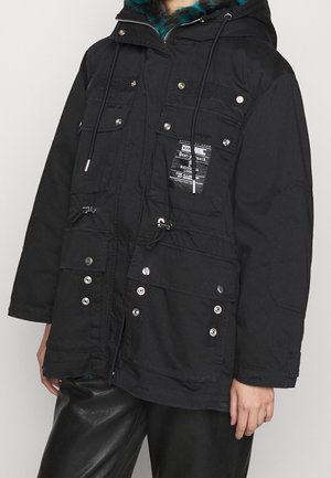 Parka noire avec une finition texturée, plusieurs poches avant, fermetures à pression et une capuche en fourrure. Présente une étiquette distinctive.