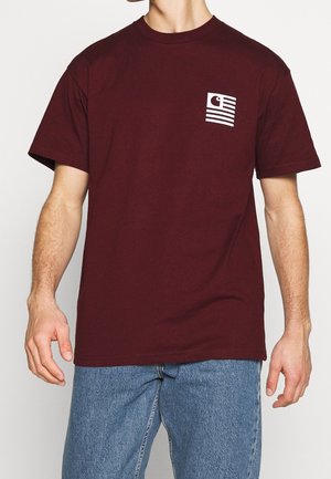 Hombre con camiseta de manga corta de color rojo oscuro con un pequeño logo blanco en el pecho, combinado con vaqueros azules.