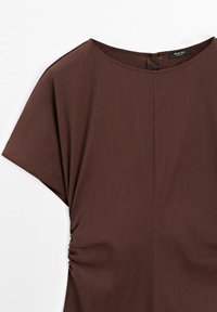 Blouse marron à manches courtes avec encolure ronde et détail froncé sur le côté, montrant le haut devant et la manche gauche sur un fond blanc.