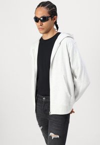 Svijetlosiva zip-up hoodie s kapuljačom, uparen s crnom majicom i crnim uništenim trapericama, s glatkom teksturom i prednjim džepovima.